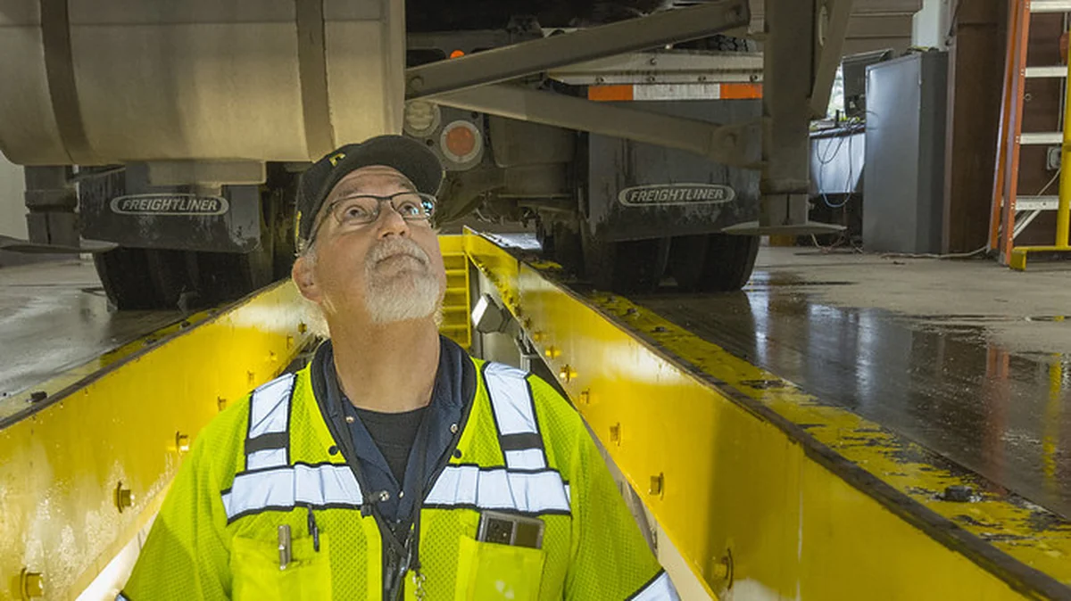 DOT inspector in high-visibility vest inspecting the underside of a truck at an inspection station